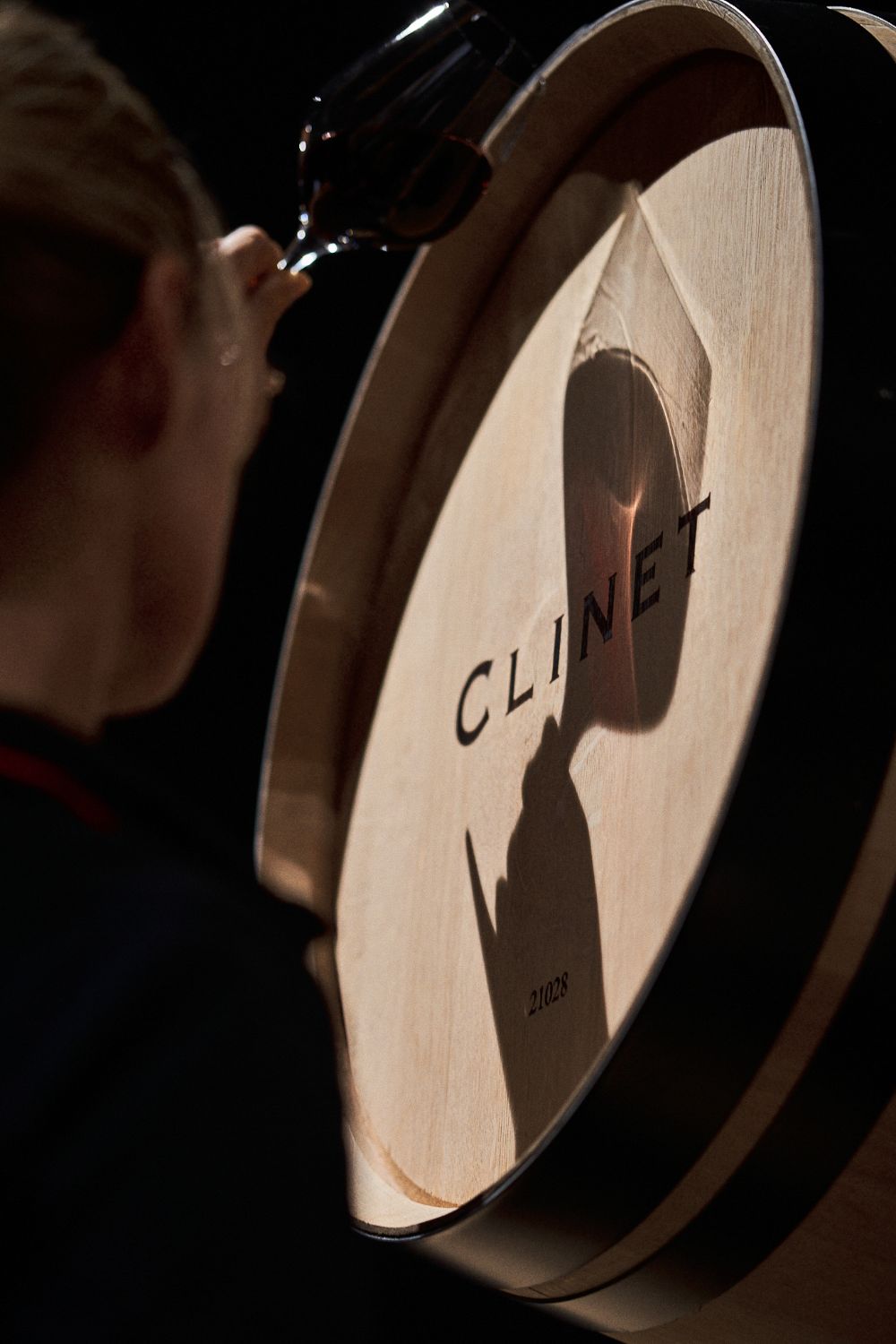 A woman tasting wine in front of a wooden Clinet barrel, projecting shadows onto the barrel.